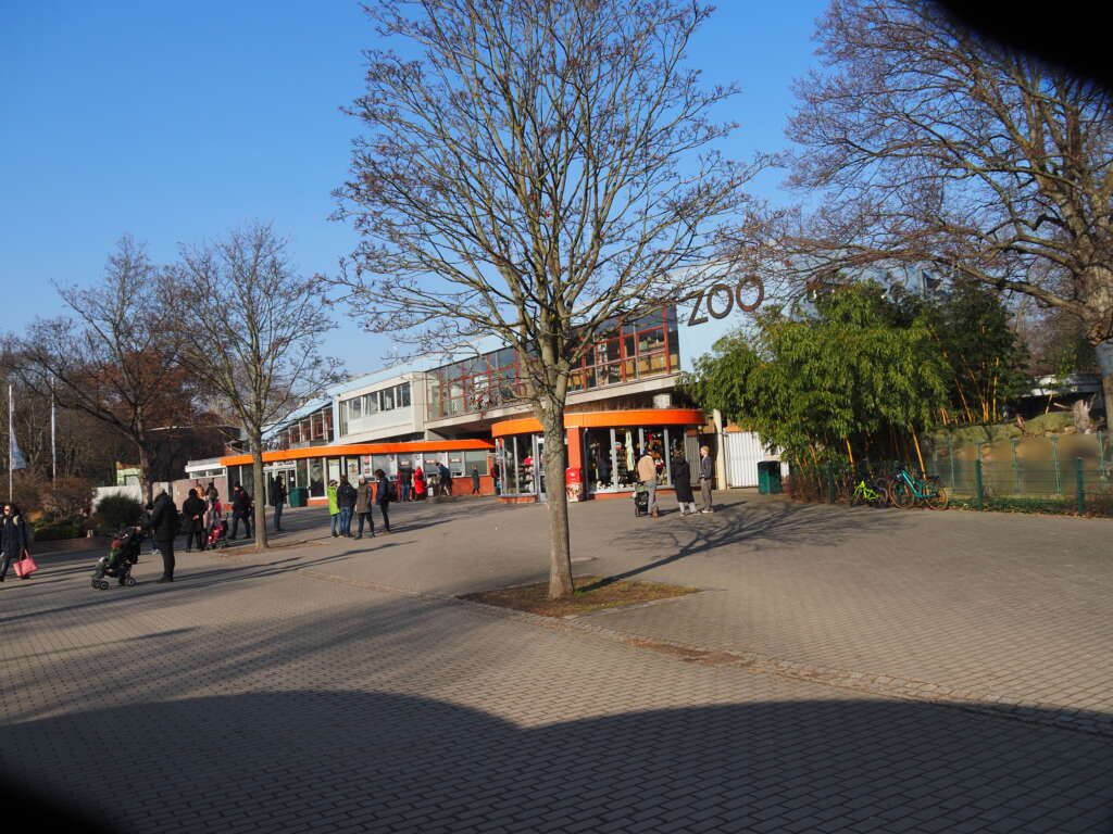 Eingangsbereich eines Zoos mit orangefarbenen Vordächern, kahlen Bäumen und Besuchern an einem sonnigen Wintertag.