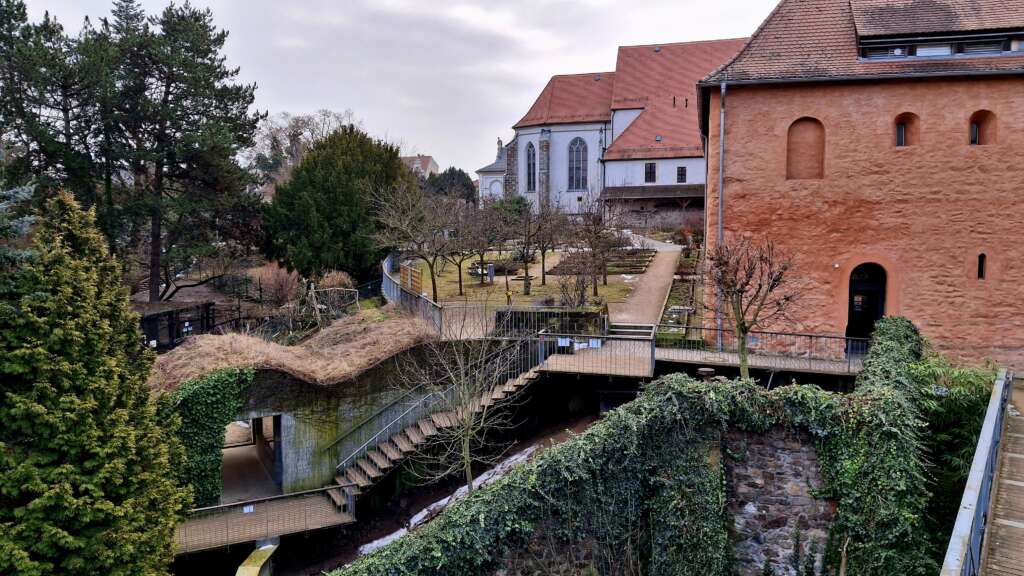 Mittelalterliche Klosteranlage mit rotem Sandsteinturm, Steganlage, Gartenbereich und gotischer Kirche im Hintergrund