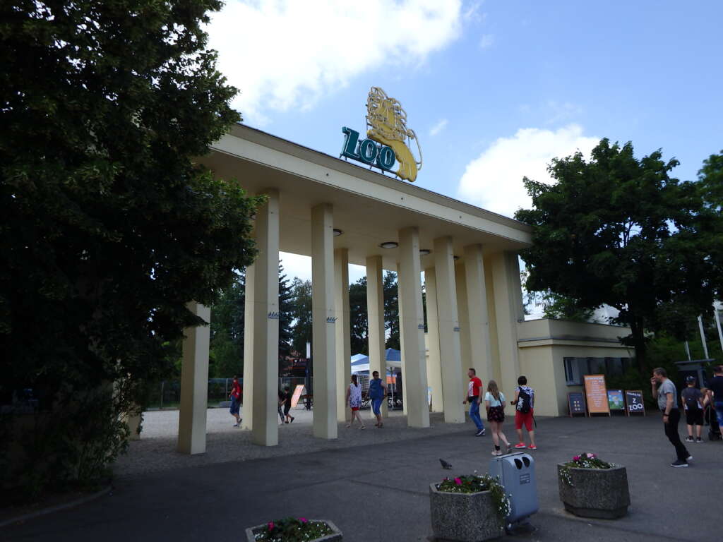 Eingangsportal eines Zoos mit Säulen und goldenem Löwen-Logo auf dem Dach, Besucher davor